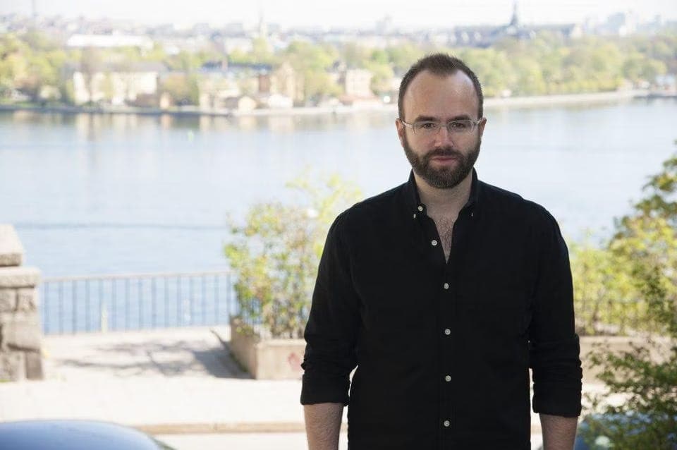 Magnus Larsson, a white man wearing a black shirt and glasses, standing in front of a lake during the day.